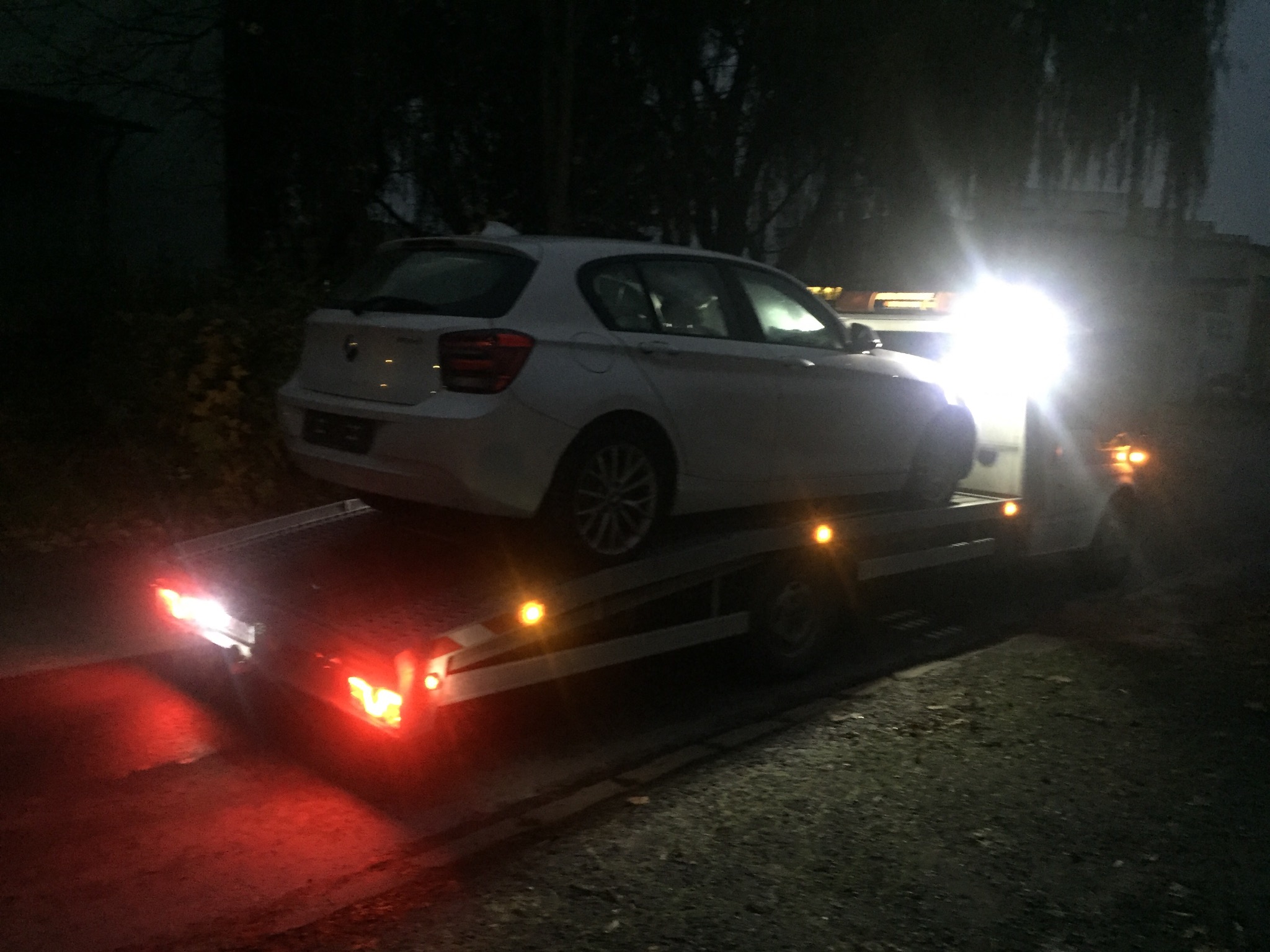 Biały samochód osobowy BMW serii 1 załadowany na platformę lawety w nocy, oświetlony światłami lawety.