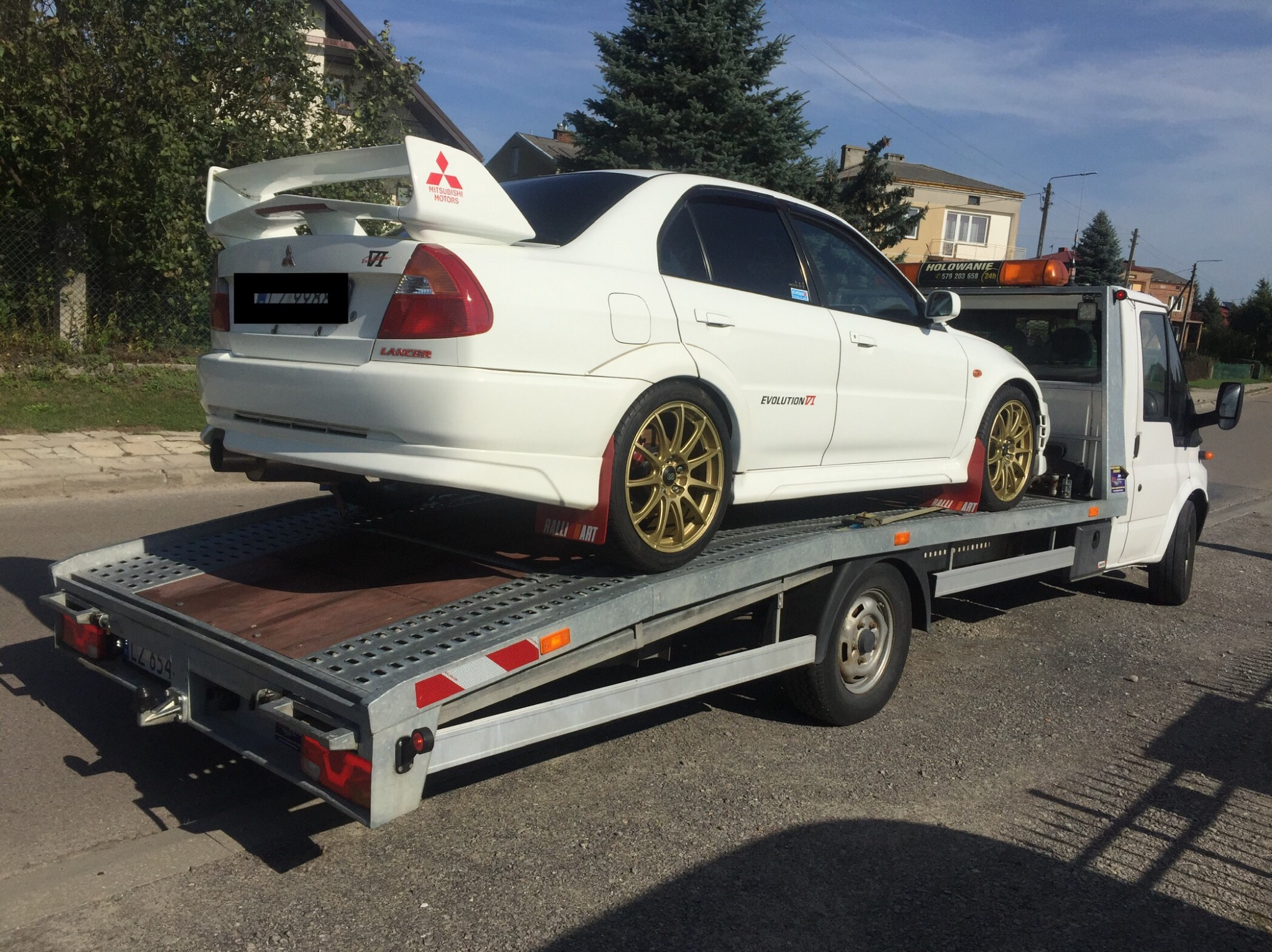 Biały sportowy samochód Mitsubishi Lancer Evolution VI z dużym spojlerem, złotymi felgami i naklejkami Ralliart umieszczony na platformie lawety samochodowej z włączonymi pomarańczowymi światłami...