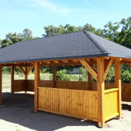 Drewniana altana z dachem pokrytym gontem bitumicznym, z niskimi ściankami, otoczona zielenią w słoneczny dzień.