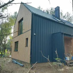 Nowoczesny dom z drewnianą elewacją i blachą na dachu, otoczony drzewami, z rurami PCV leżącymi na ziemi.