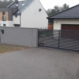 Nowoczesna posesja z przesuwną bramą poziomą z szarych profili, osadzoną w szarym, tynkowanym murze z furtką po lewej stronie, na tle zabudowy jednorodzinnej.