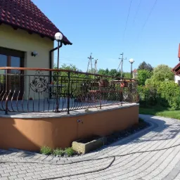 Kuty balkon z drewnianą poręczą, zdobiony motywami roślinnymi, przylegający do domu z czerwoną dach&oacute;wką, widok z poziomu kostki brukowej z wężem ogrodowym.