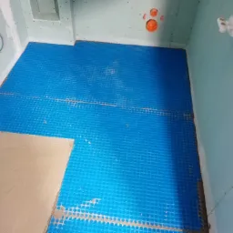 Niebieska mata grzewcza pod płytki, częściowo pokryta wylewką samopoziomującą, w surowej łazience z instalacją prysznicową i odpływem liniowym w podłodze.