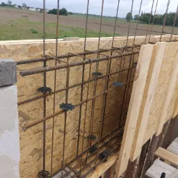 Zbrojenie pionowe i poziome w szalunku z płyty OSB, przygotowane do zalania betonem, widoczne dystanse z tworzywa sztucznego, w tle pole uprawne i zabudowania.