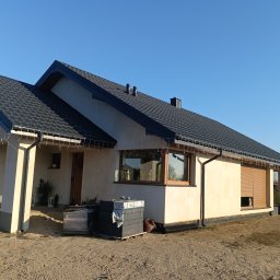 Baubud Michał Przebieracz Usługi wykonawcze i projektowe - Nowoczesny dom jednorodzinny z ciemną dachówką i beżową elewacją, w trakcie wykończenia terenu wokół budynku. Widoczne okna z roletami zewnętrznymi i dekoracyjne oświetlenie.