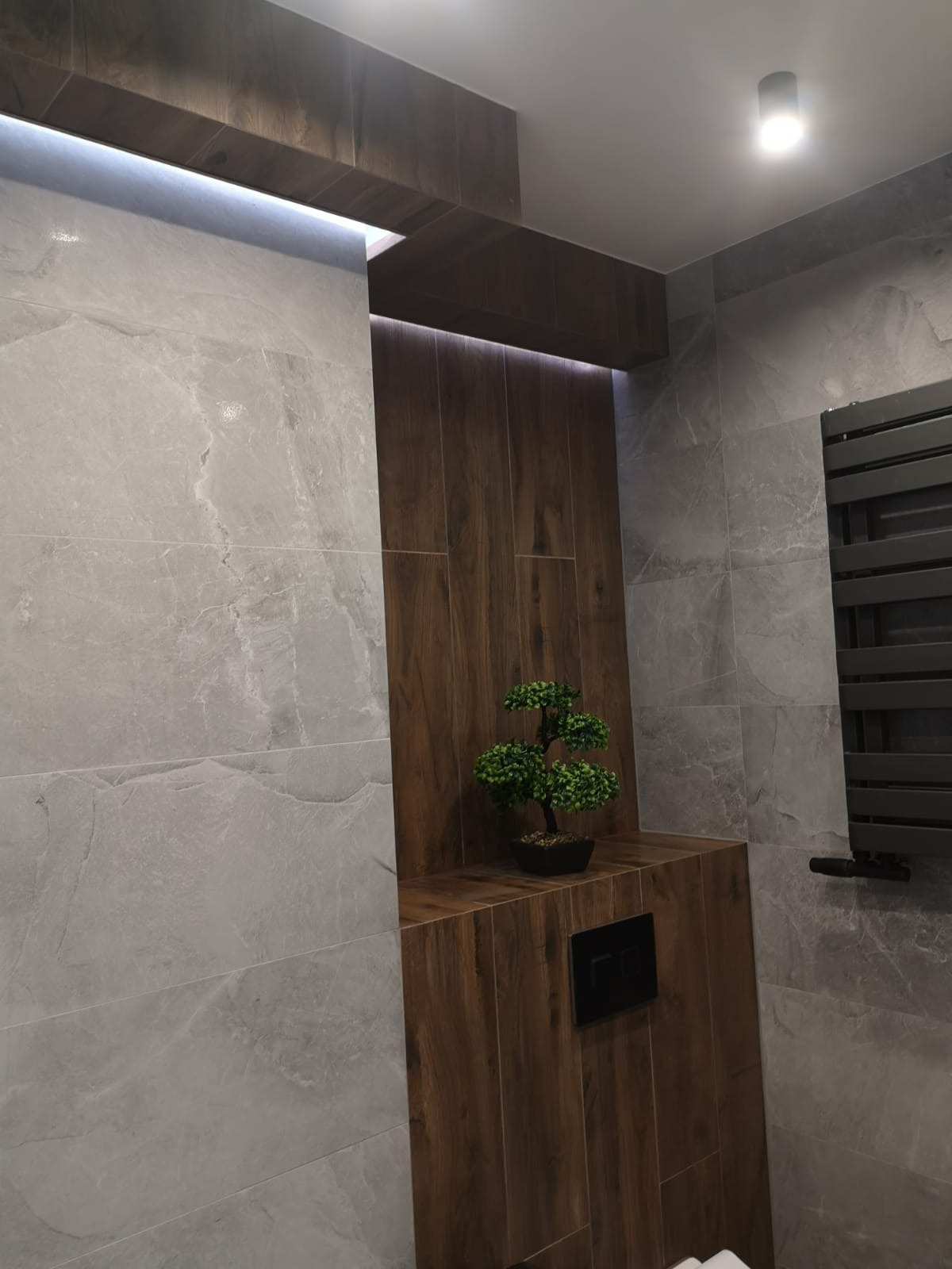 Nowoczesna łazienka z szarymi płytkami imitującymi marmur, drewnopodobną wnęką z podświetleniem LED i dekoracyjnym drzewkiem bonsai, z widocznym grzejnikiem drabinkowym.