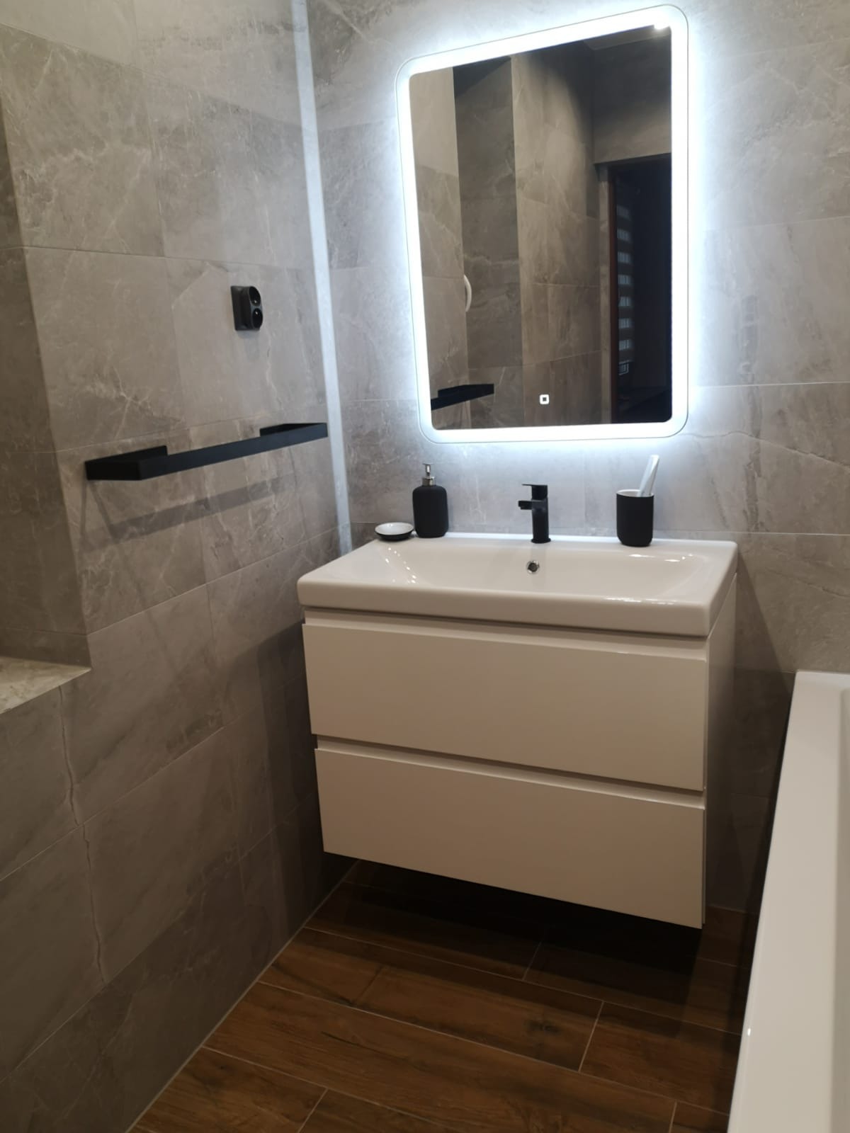 Nowoczesna łazienka z szarymi płytkami imitującymi marmur, drewnopodobną podłogą, białą szafką pod umywalkę z czarną armaturą i podświetlanym lustrem LED.