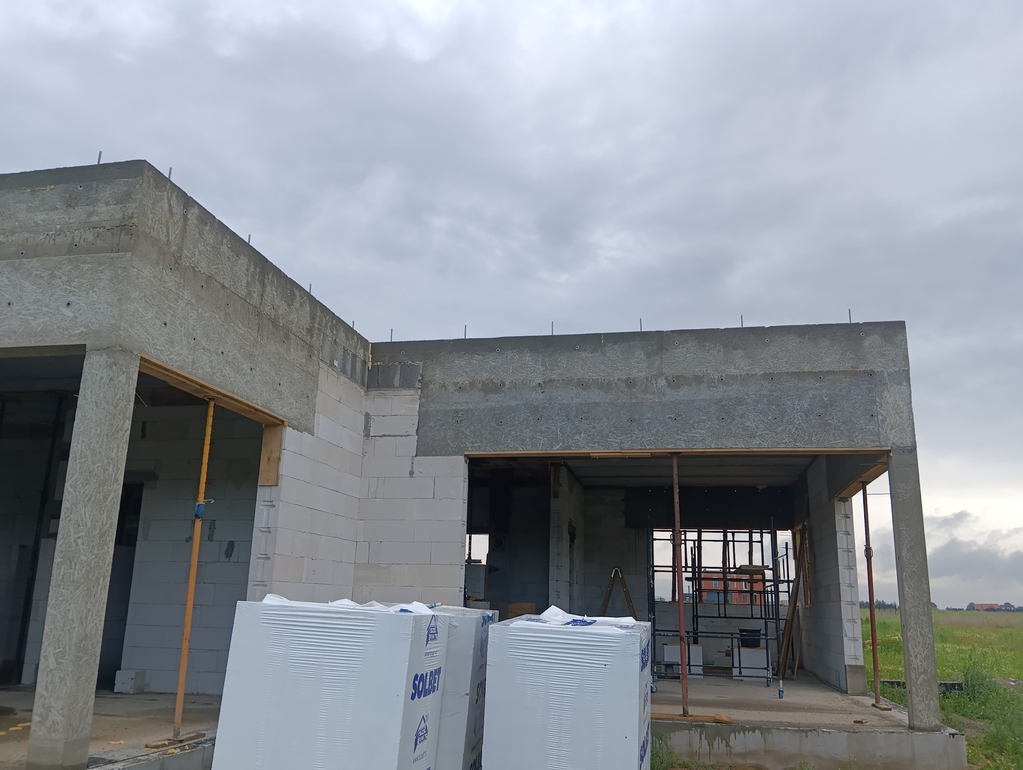 Surowy stan domu z betonu komórkowego w trakcie budowy, widoczne podpory, szalunki i materiały budowlane Solbet na pierwszym planie, pochmurne niebo w tle.