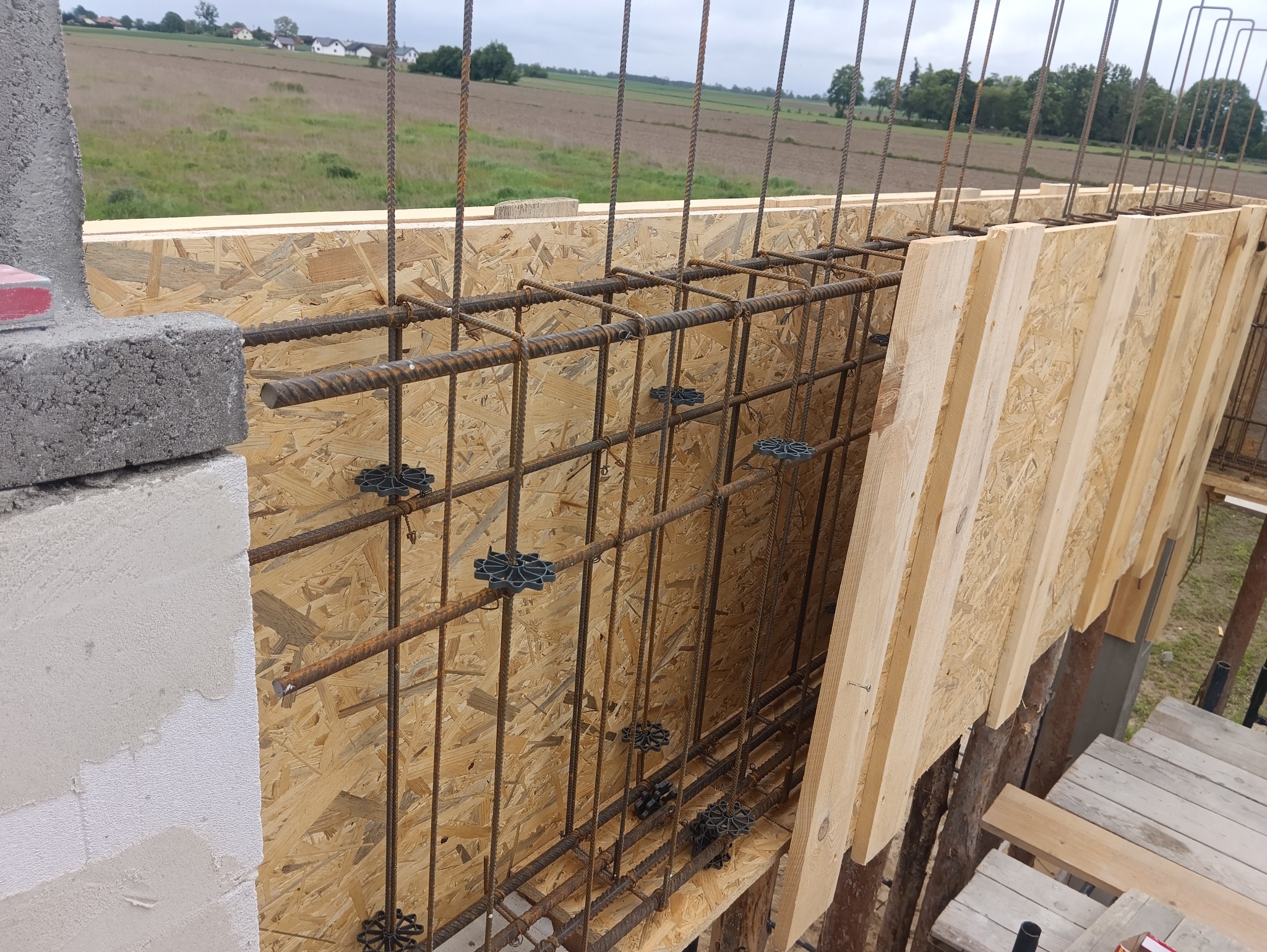 Zbrojenie pionowe i poziome w szalunku z płyty OSB, przygotowane do zalania betonem, widoczne dystanse z tworzywa sztucznego, w tle pole uprawne i zabudowania.
