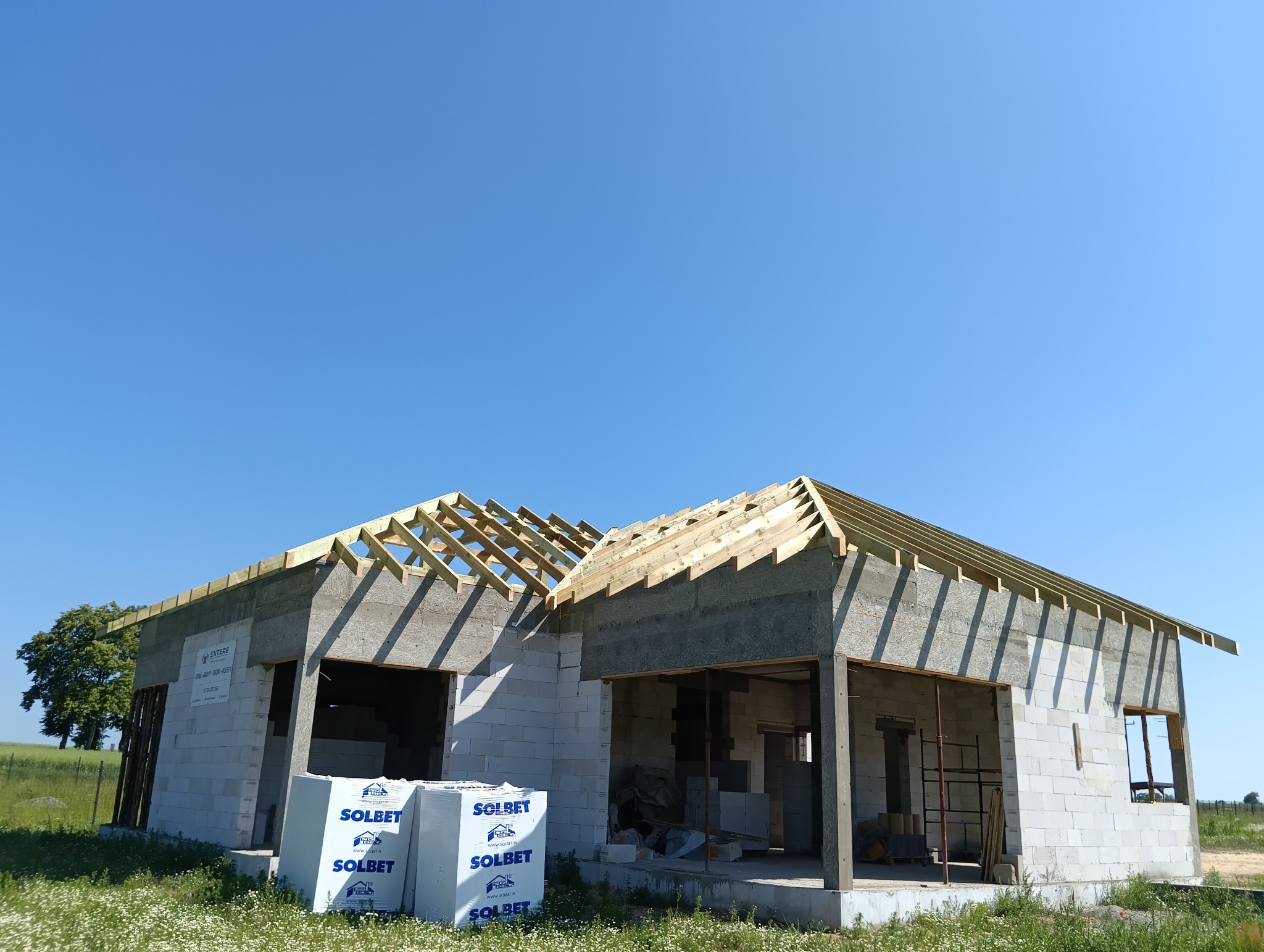 Budowa domu jednorodzinnego w stanie surowym otwartym: ściany z betonu komórkowego, drewniana konstrukcja dachu, palety bloczków Solbet na pierwszym planie, błękitne niebo w tle.
