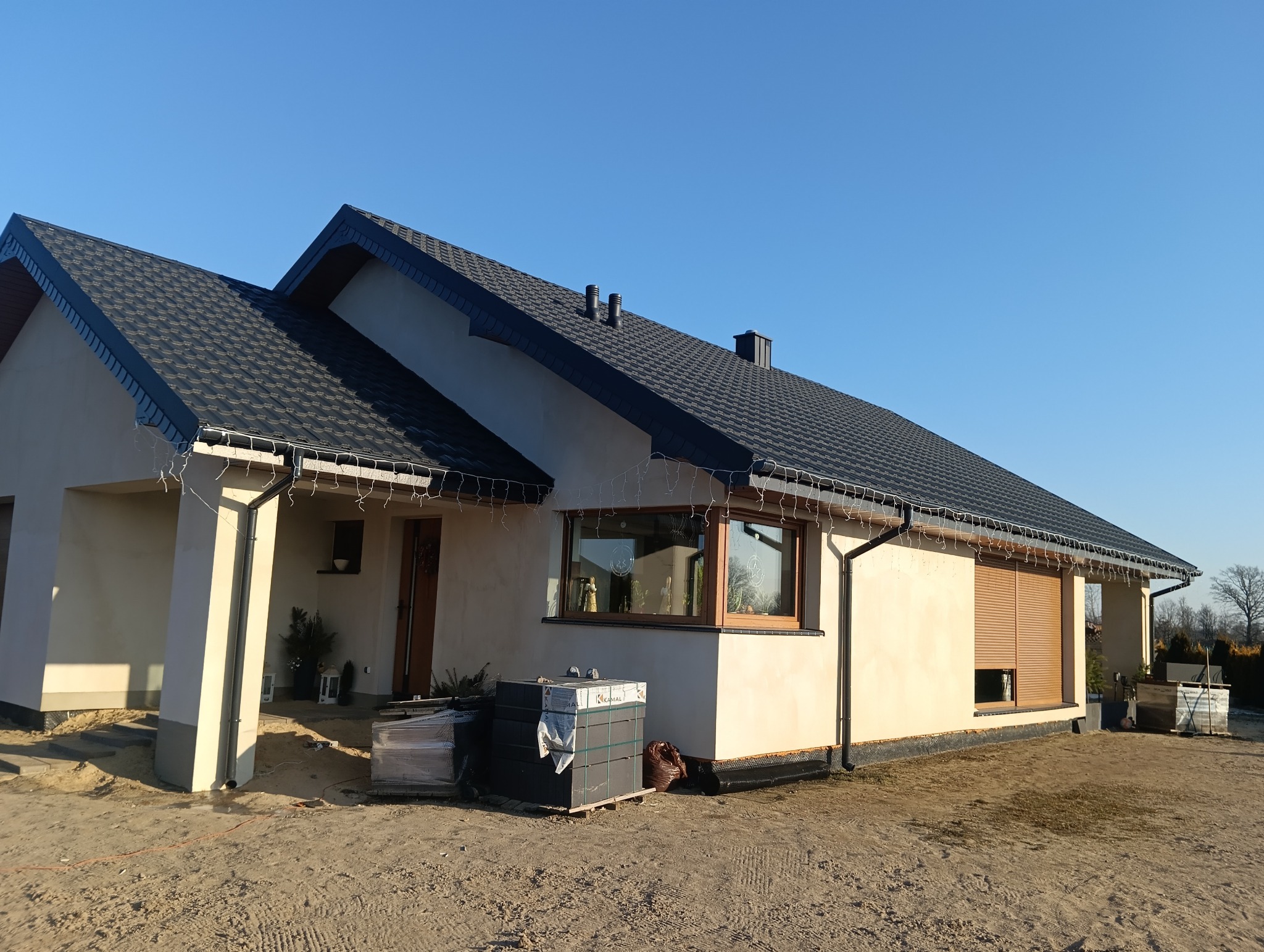 Nowoczesny dom jednorodzinny z ciemną dachówką i beżową elewacją, w trakcie wykończenia terenu wokół budynku. Widoczne okna z roletami zewnętrznymi i dekoracyjne oświetlenie.