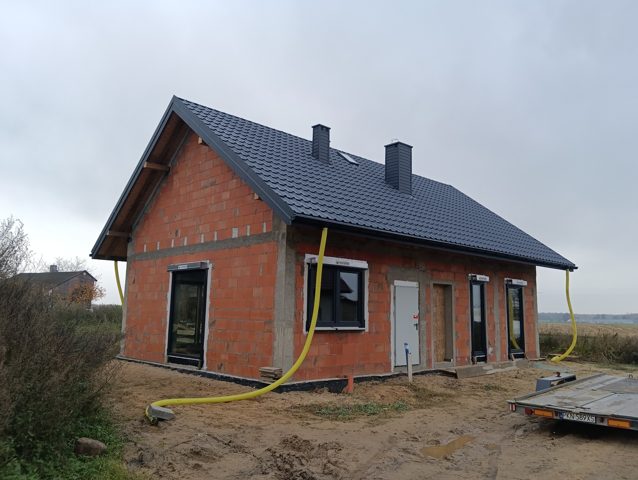 Surowy dom z czerwonej cegły z czarnym dachem i oknami, widoczne rury wentylacyjne, teren budowy. Budynek w trakcie budowy, widok z zewnątrz.