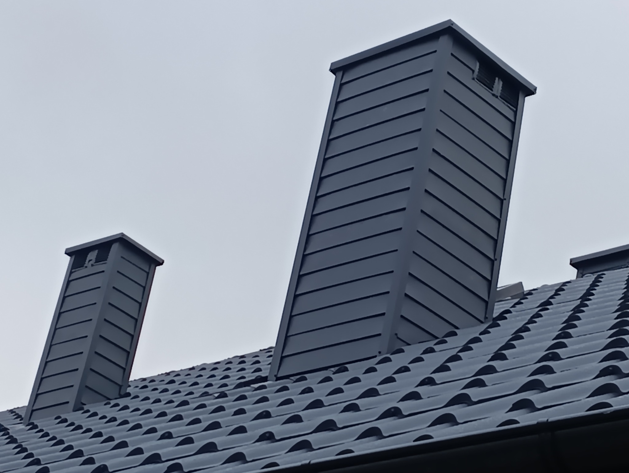 Grafitowy dach z dachówki ceramicznej z dwoma kominami o nowoczesnej, prostej formie. Jeden komin jest wyższy od drugiego. Pochmurne niebo w tle.