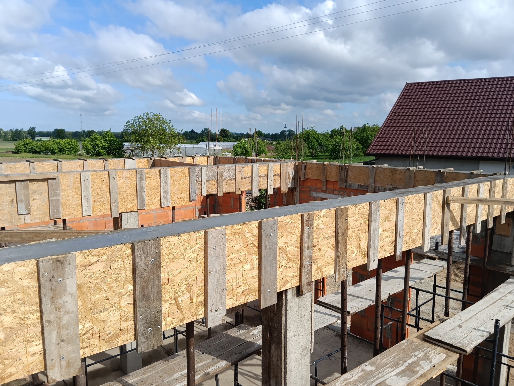 Fundamenty domu w trakcie budowy: drewniane szalunki, cegły i stalowe pręty zbrojeniowe na tle błękitnego nieba z chmurami. Widok z góry.