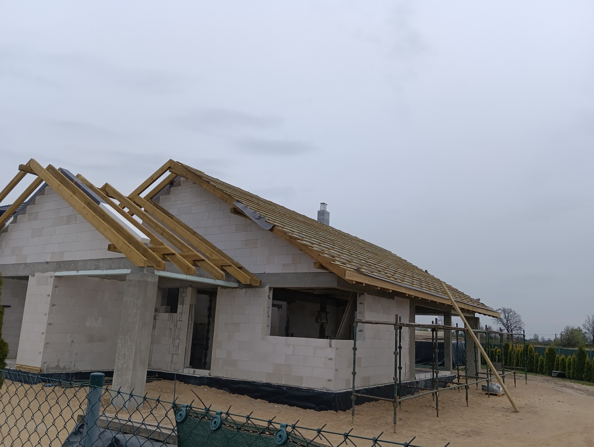 Budowa domu z białych bloczków: widoczne ściany, drewniana konstrukcja dachu w trakcie montażu, komin. Ujęcie z dołu, ogrodzenie w tle.