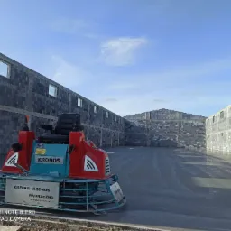 Zacieraczka mechaniczna do betonu marki Kronos na tle surowych ścian hali w budowie w Ostrowie Wielkopolskim. Świeżo wylana, gładka posadzka betonowa odbija światło słoneczne.