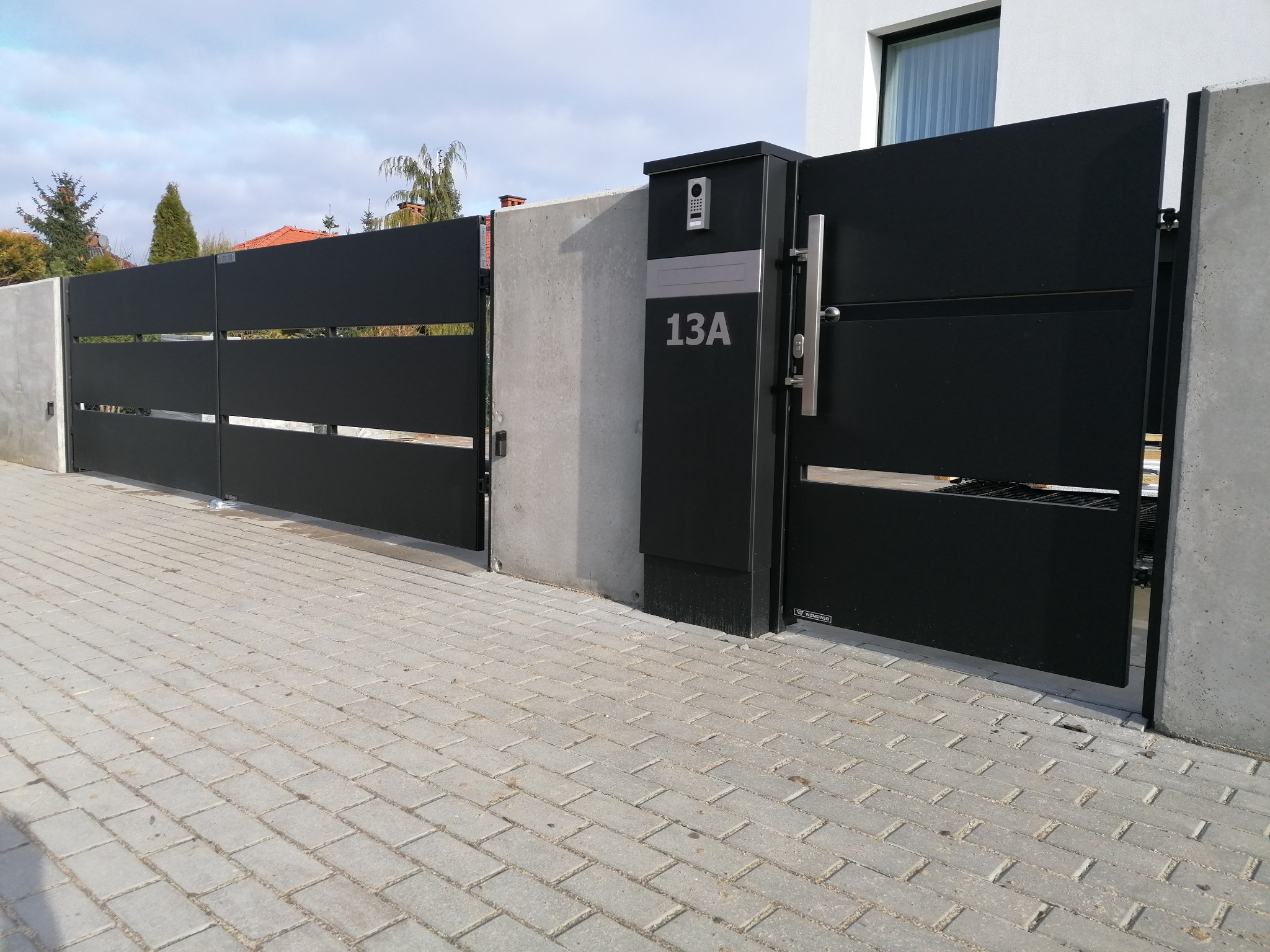 Nowoczesna, przesuwna brama wjazdowa i furtka z numerem domu 13A, w kolorze czarnym, zamontowane na betonowych słupkach i kostce brukowej.