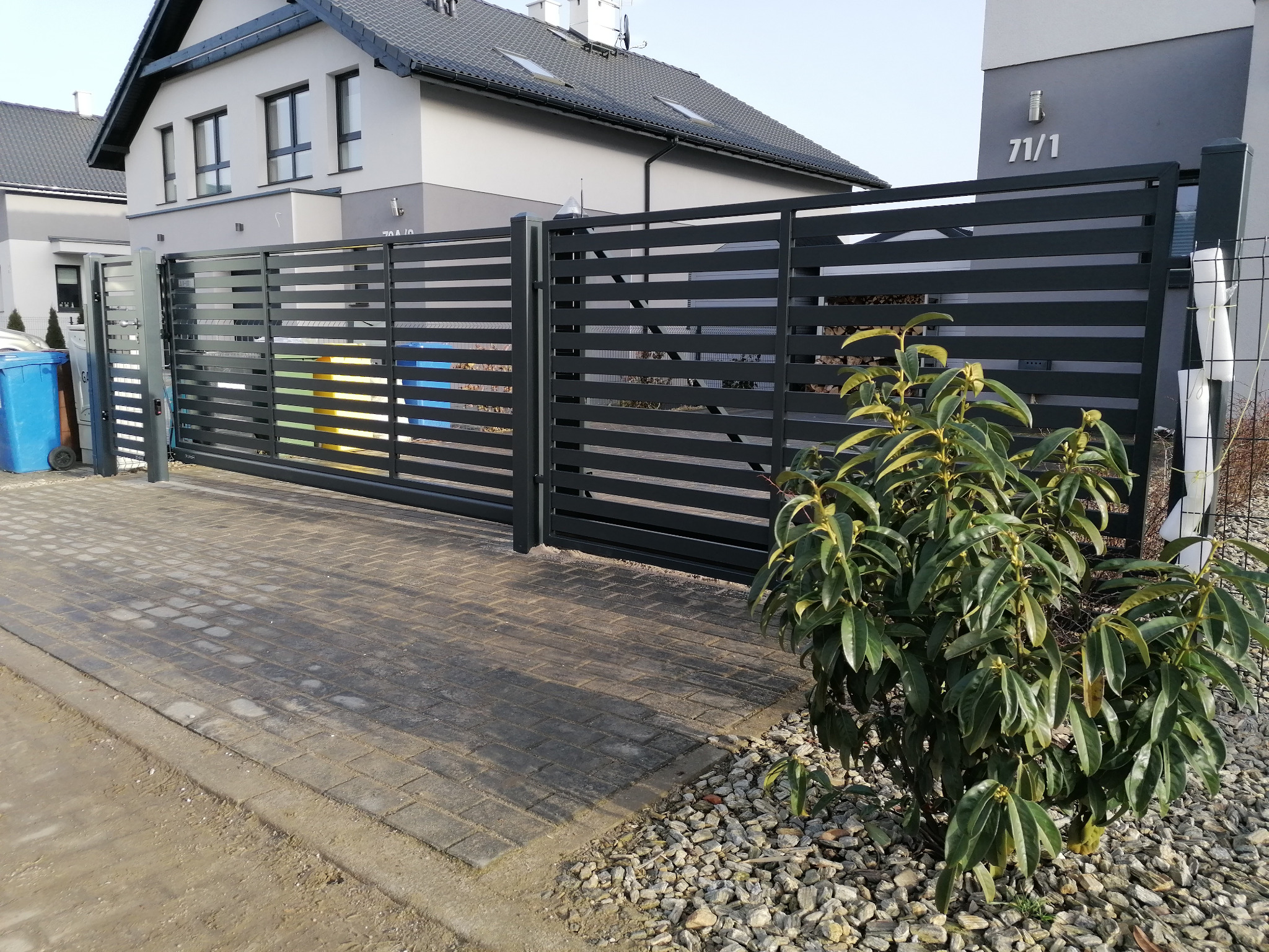 Nowoczesna, pozioma brama wjazdowa i przesuwna, w kolorze antracytowym, na tle nowego domu jednorodzinnego z numerem 71/1, z kostki brukowej i ozdobnego krzewu.