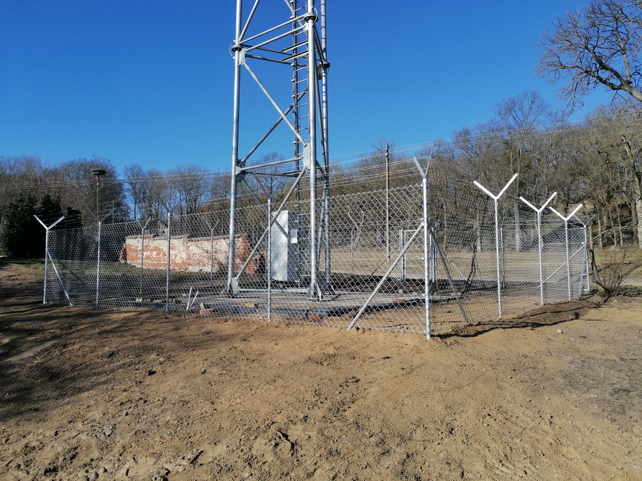 Nowo postawione ogrodzenie z siatki z drutem kolczastym otaczające stalową wieżę telekomunikacyjną na tle błękitnego nieba i drzew bez liści.