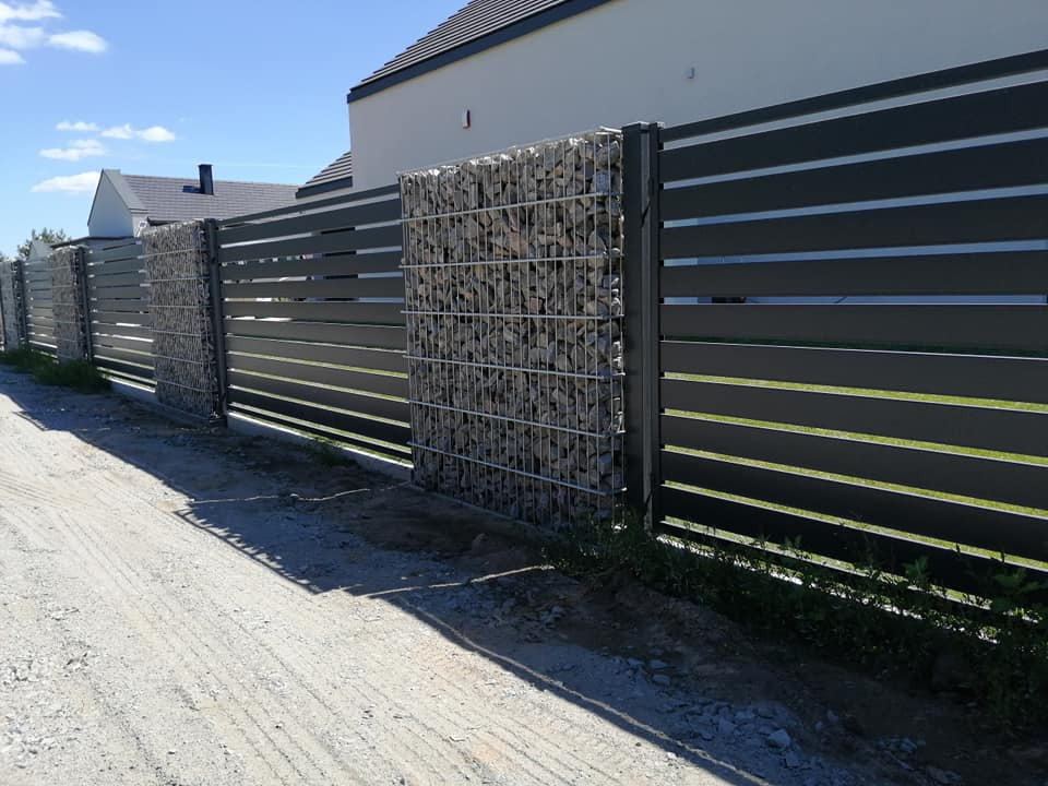 Nowoczesne ogrodzenie z poziomych, grafitowych paneli z gabionowymi słupkami wypełnionymi szarymi kamieniami, na tle domu z ciemną dachówką i błękitnego nieba.