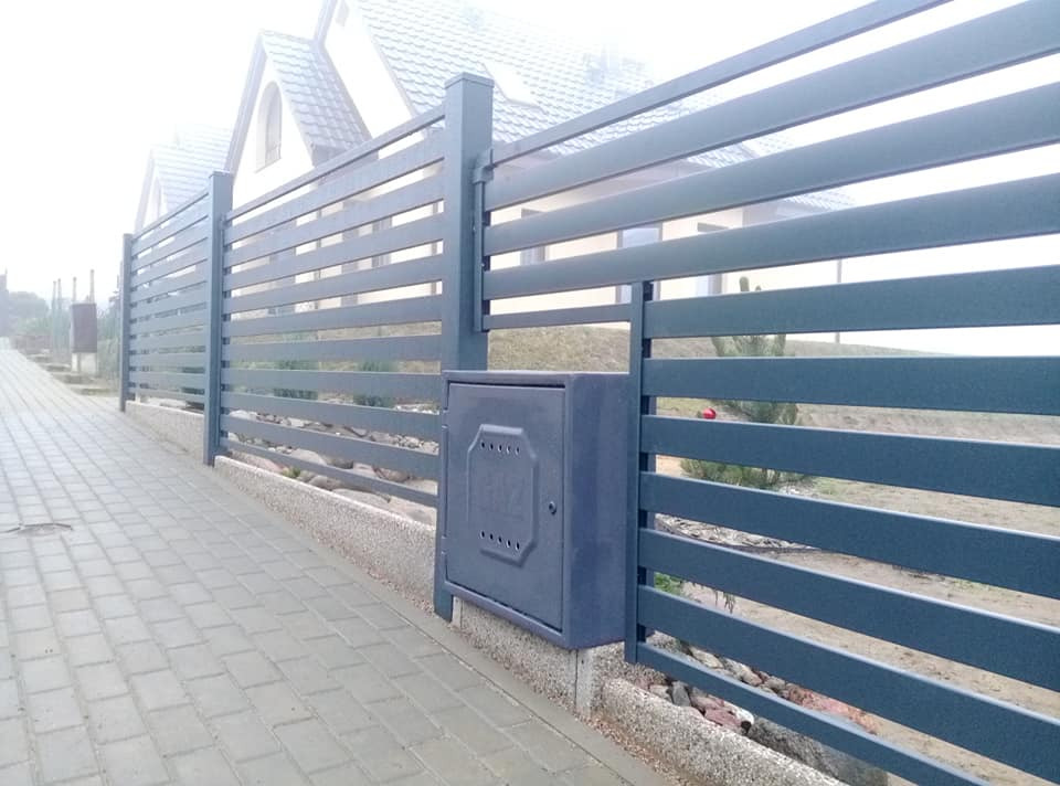 Szary, poziomy płot z metalowych paneli z zamontowaną skrzynką na gaz przy chodniku z kostki brukowej, w tle dom we mgle.