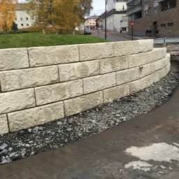 Niski, łukowaty murek oporowy z jasnych, prostokątnych bloków kamiennych, z podsypką z ciemnego żwiru, na tle miejskiego krajobrazu z budynkami i drzewami o jesiennych liściach.