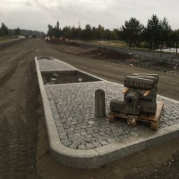 Bruk-Majstero - Układanie kostki granitowej na placu budowy, widoczne palety z kostką, częściowo ułożona nawierzchnia z obrzeżami, w tle ciężki sprzęt i krajobraz z drzewami pod pochmurnym niebem.