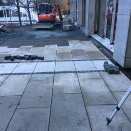 Bruk-Majstero - Trwa układanie dużych, szarych płyt tarasowych przy budynku z szarej cegły. Na tyłach widoczna koparka i przygotowane kostki. W kadrze fragment niwelatora.