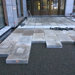 Bruk-Majstero - Montaż tarasu z płyt kamiennych na przygotowanym podłożu, widoczny poziomica i małe kostki do wykończenia krawędzi, w tle budynek z dużymi oknami.