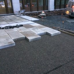 Bruk-Majstero - Fragment budowy tarasu: ułożone płyty kamienne na podsypce, w tle budynek z wózkiem budowlanym i zaparkowany samochód dostawczy.