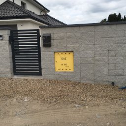 Bruk-Majstero - Nowoczesne ogrodzenie z szarej kostki elewacyjnej z czarną skrzynką na listy i metalową bramką, widoczny żółty zawór gazowy i teren przygotowany pod dalsze prace.