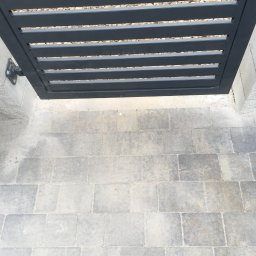 Bruk-Majstero - Fragment nowoczesnego ogrodzenia panelowego w kolorze antracytowym z poziomymi szczeblami, widok z góry na szarą kostkę brukową i drobne kamyki dekoracyjne w tle.