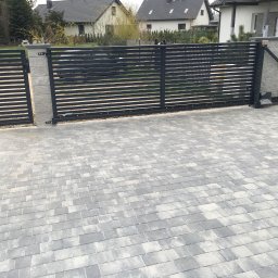 Bruk-Majstero - Nowoczesna, pozioma, metalowa brama wjazdowa z automatyką, osadzona na szarych, betonowych słupkach, na tle ułożonej szarej kostki brukowej i domów jednorodzinnych.