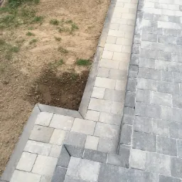 Świeżo ułożona nawierzchnia z kostki brukowej w odcieniach szarości i beżu, obrzeża z ciemniejszej kostki, widoczny fragment terenu z ziemią i trawą.