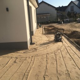Bruk-Majstero - Przygotowanie podłoża pod kostkę brukową: ubijanie piasku zagęszczarką wzdłuż obrzeży, widoczne ślady butów i niedokończone wykopy.
