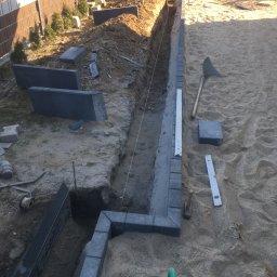 Bruk-Majstero - Przygotowany wykop pod obrzeża z ułożonymi elementami z kostki granitowej, widoczne narzędzia, sznurek wyznaczający linię, poziomica i zielona konewka na piaszczystym podłożu.