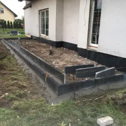 Przygotowanie podłoża pod taras: ułożone obrzeża z betonowych elementów, widoczne sznurki wyznaczające poziom, folia kubełkowa na ścianie budynku, ziemia częściowo wyrównana.