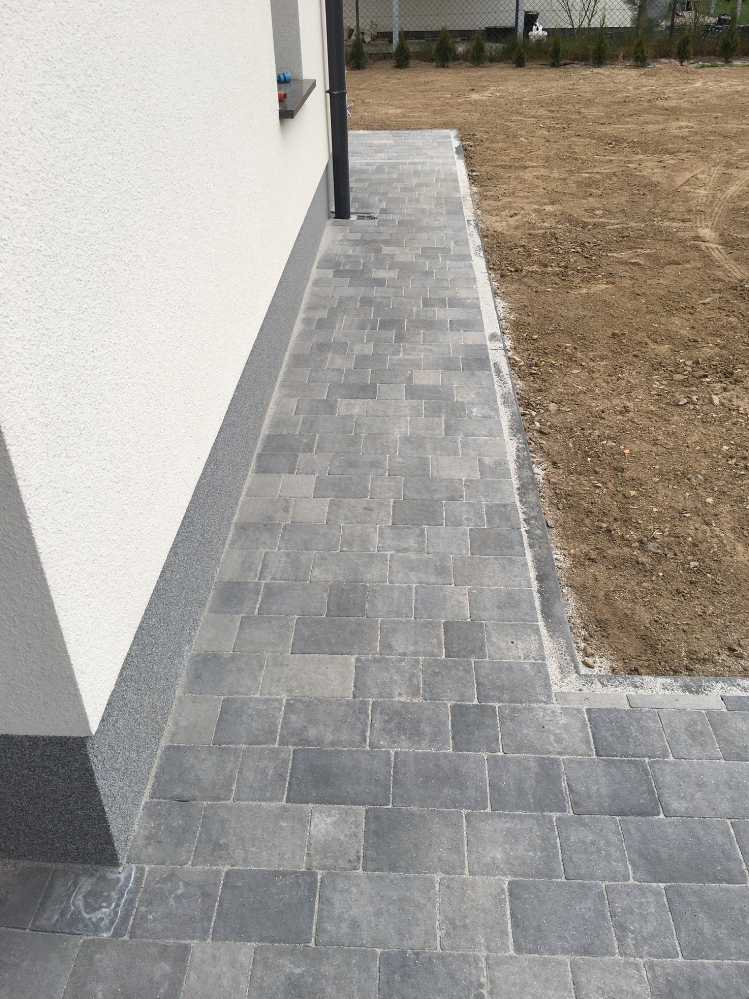 Szara ścieżka z kostki brukowej przylegająca do białej ściany budynku, widoczny fragment trawnika z przygotowanym podłożem.