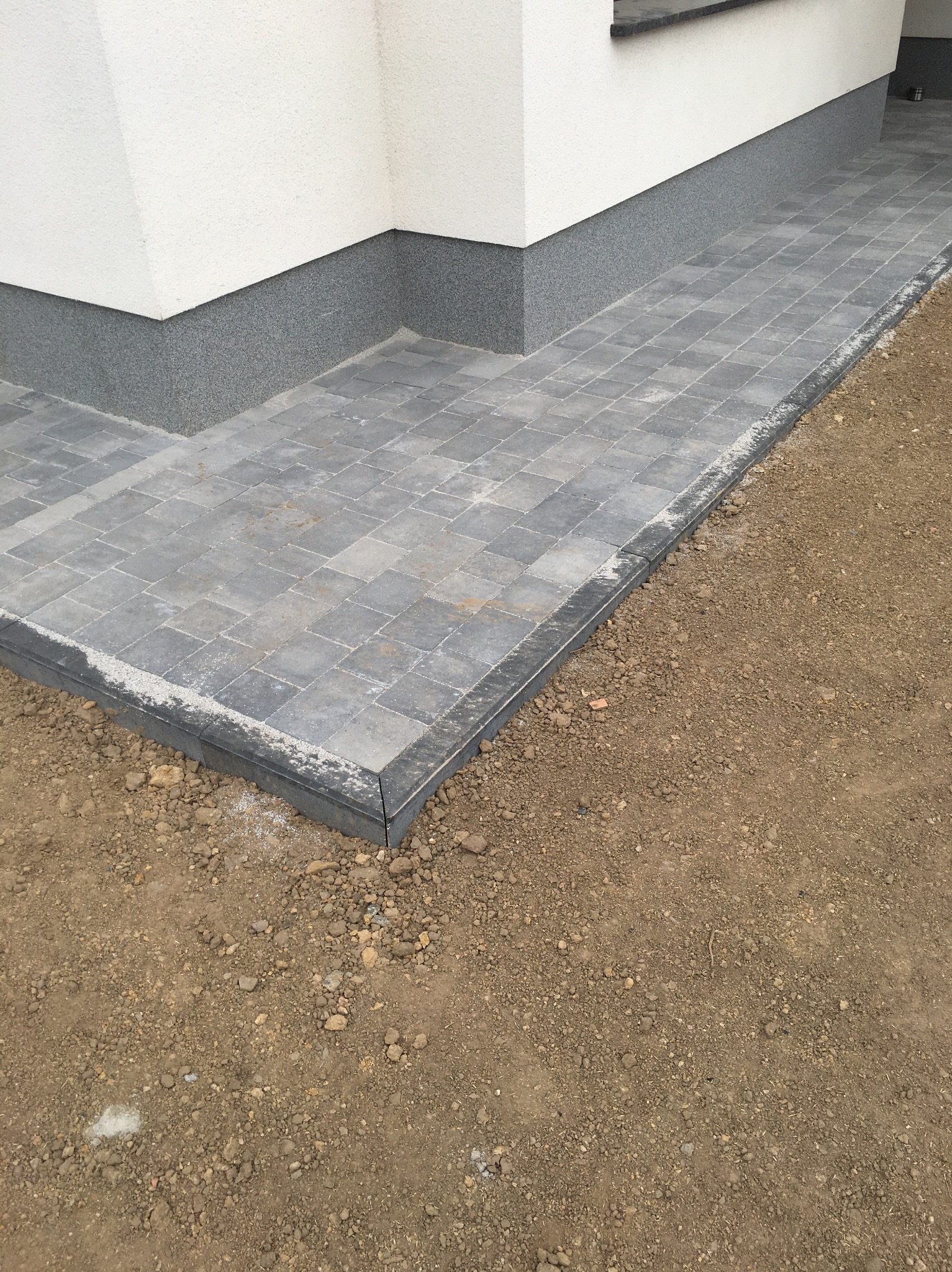 Ułożona szara kostka brukowa tworząca chodnik przy budynku z szarą elewacją, widoczne niedociągnięcia w postaci rozsypanego piasku i ziemi po bokach.
