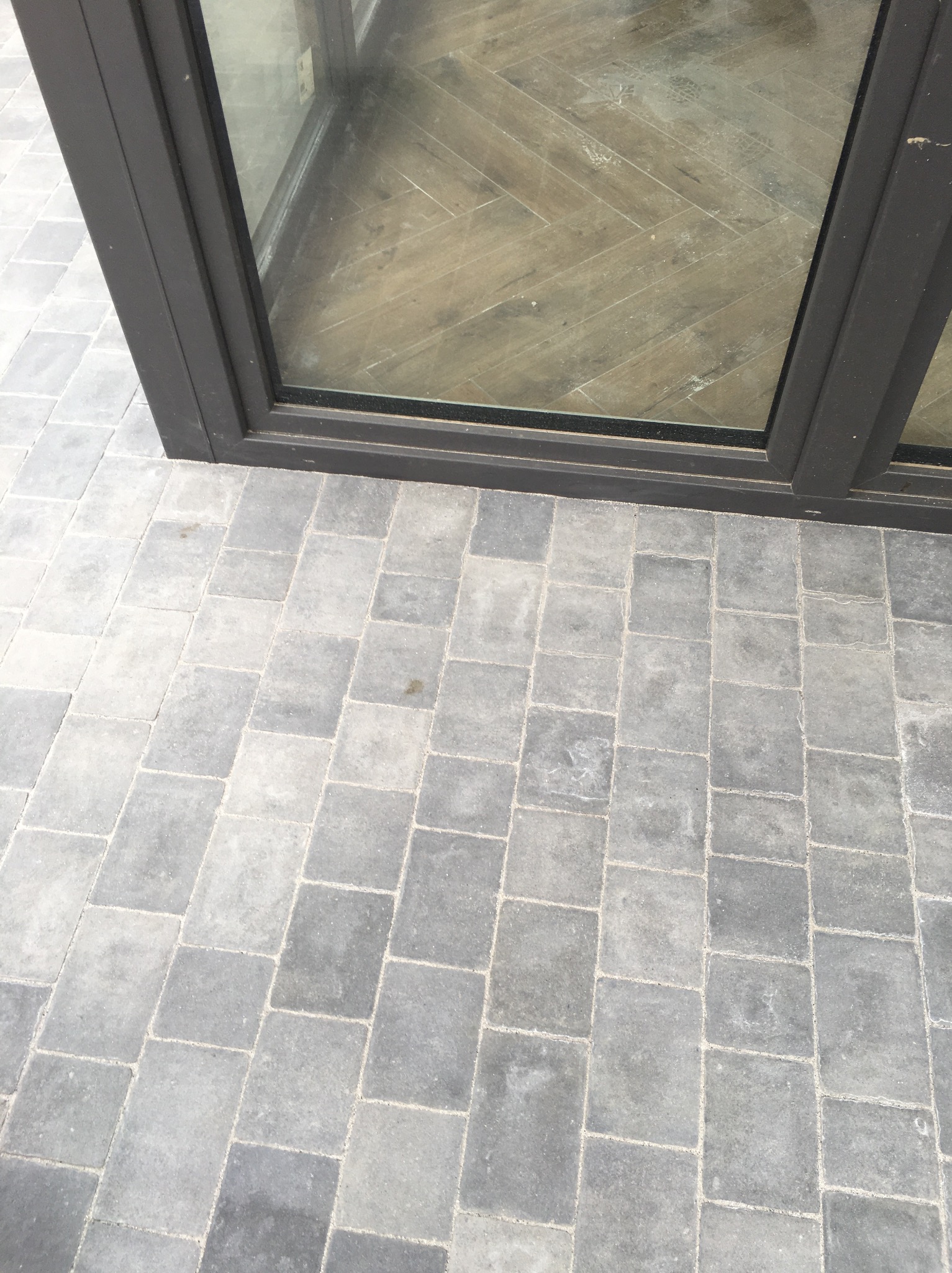 Szara kostka brukowa ułożona przy ciemnej ramie okiennej, widoczny wzór jodełki na podłodze wewnątrz budynku przez szybę.