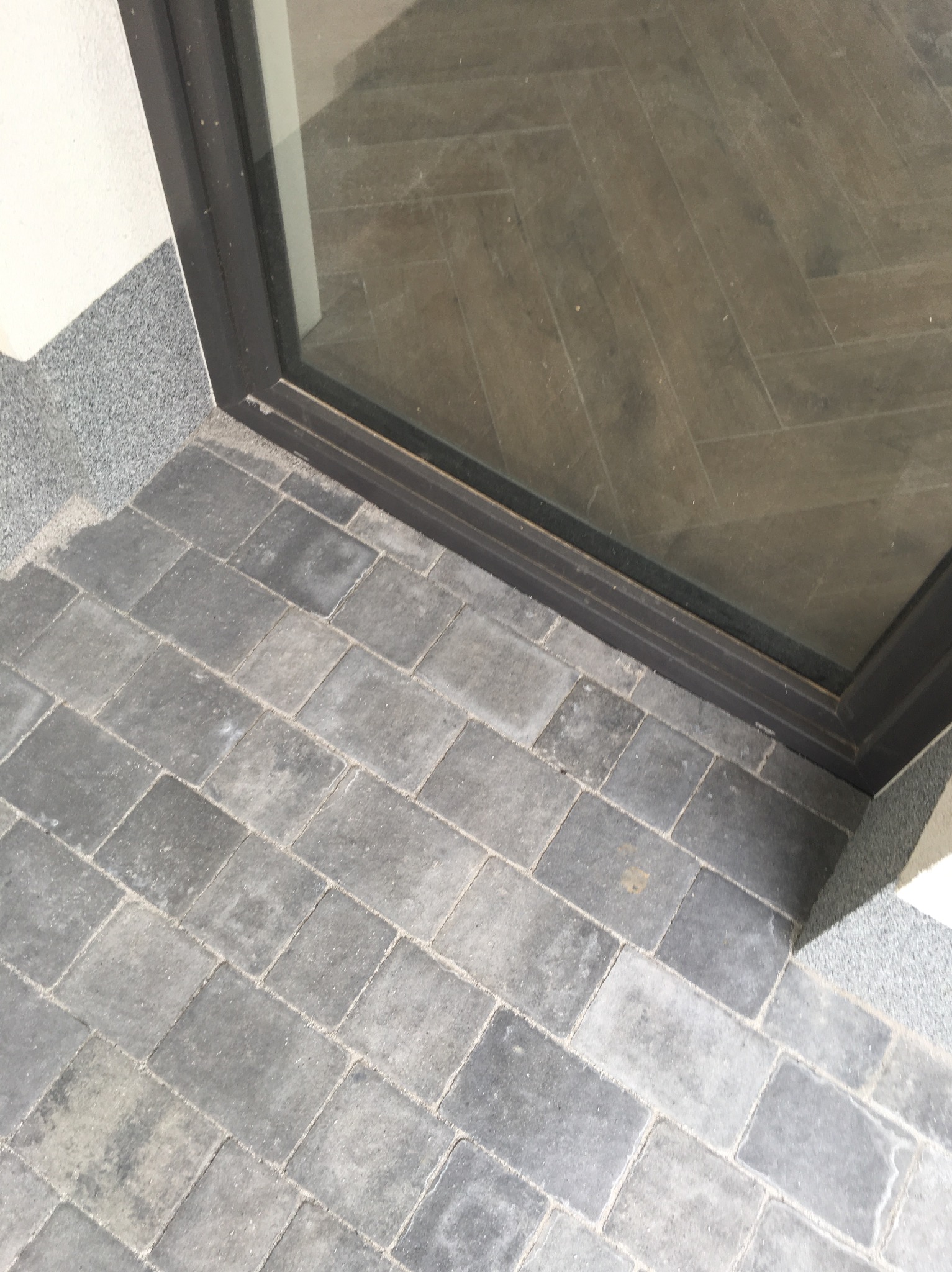 Szara kostka brukowa ułożona przy progu drzwi wejściowych, widoczny szary cokół i odbicie podłogi w szybie drzwi.