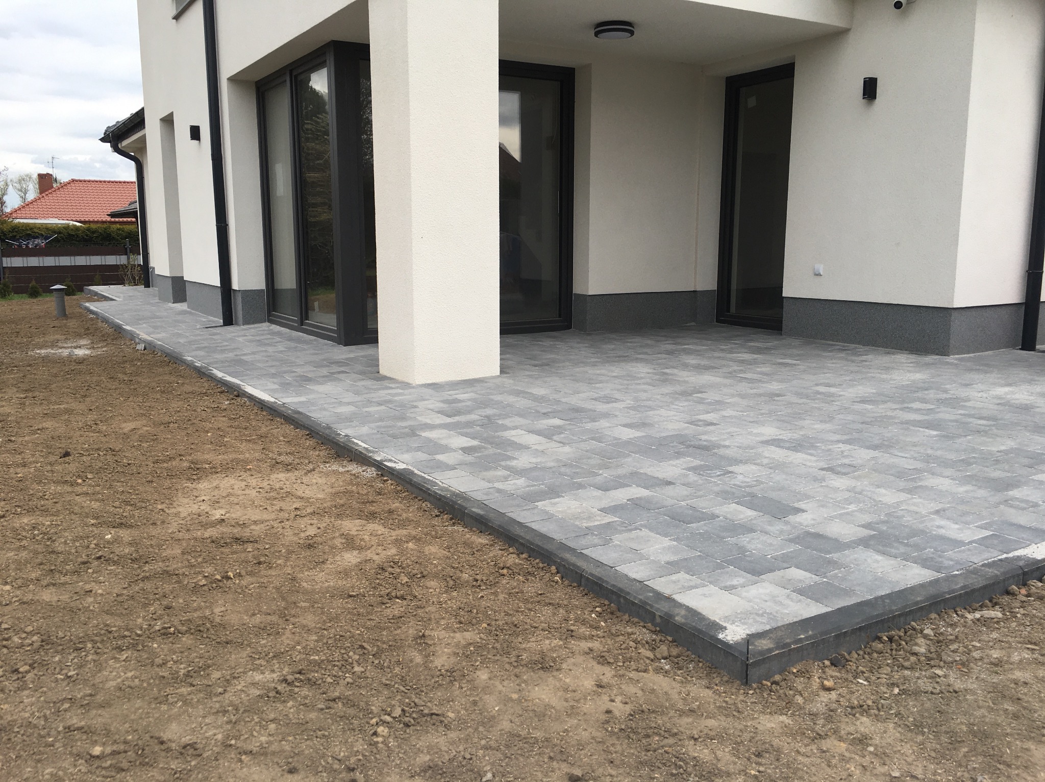 Szary taras z kostki brukowej ułożony przy nowoczesnym domu z dużymi oknami przesuwnymi, widoczny fragment niezagospodarowanego terenu.