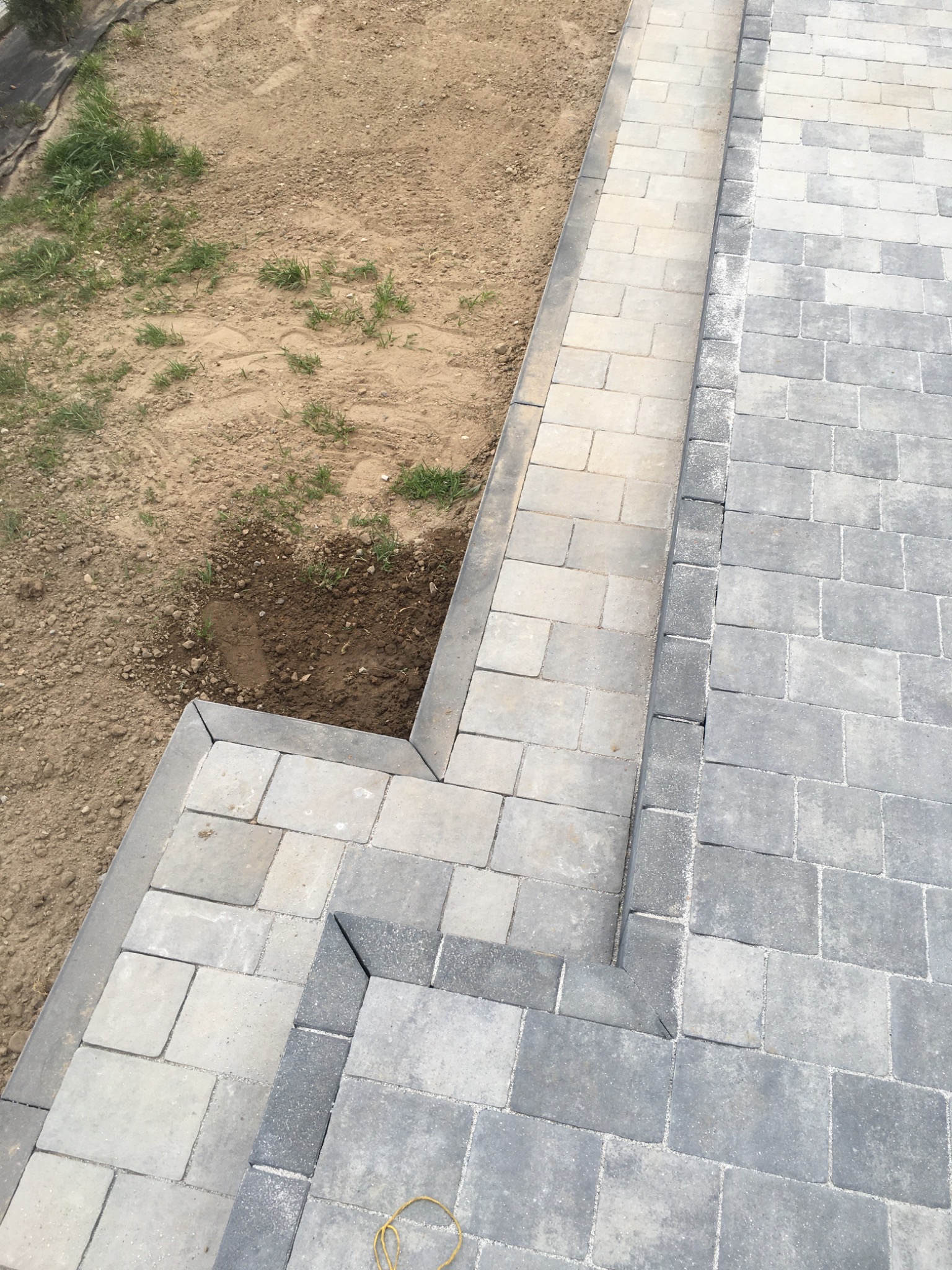 Świeżo ułożona nawierzchnia z kostki brukowej w odcieniach szarości i beżu, obrzeża z ciemniejszej kostki, widoczny fragment terenu z ziemią i trawą.