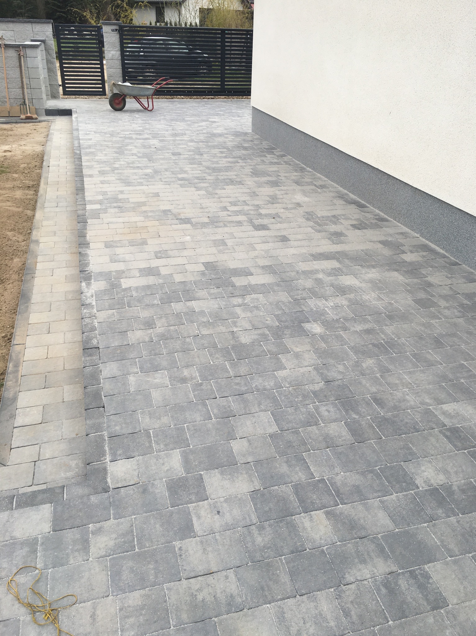 Ułożona szara kostka brukowa tworząca podjazd, widoczna taczka i fragment ogrodzenia panelowego w tle, obok ściana budynku z szarym cokołem.