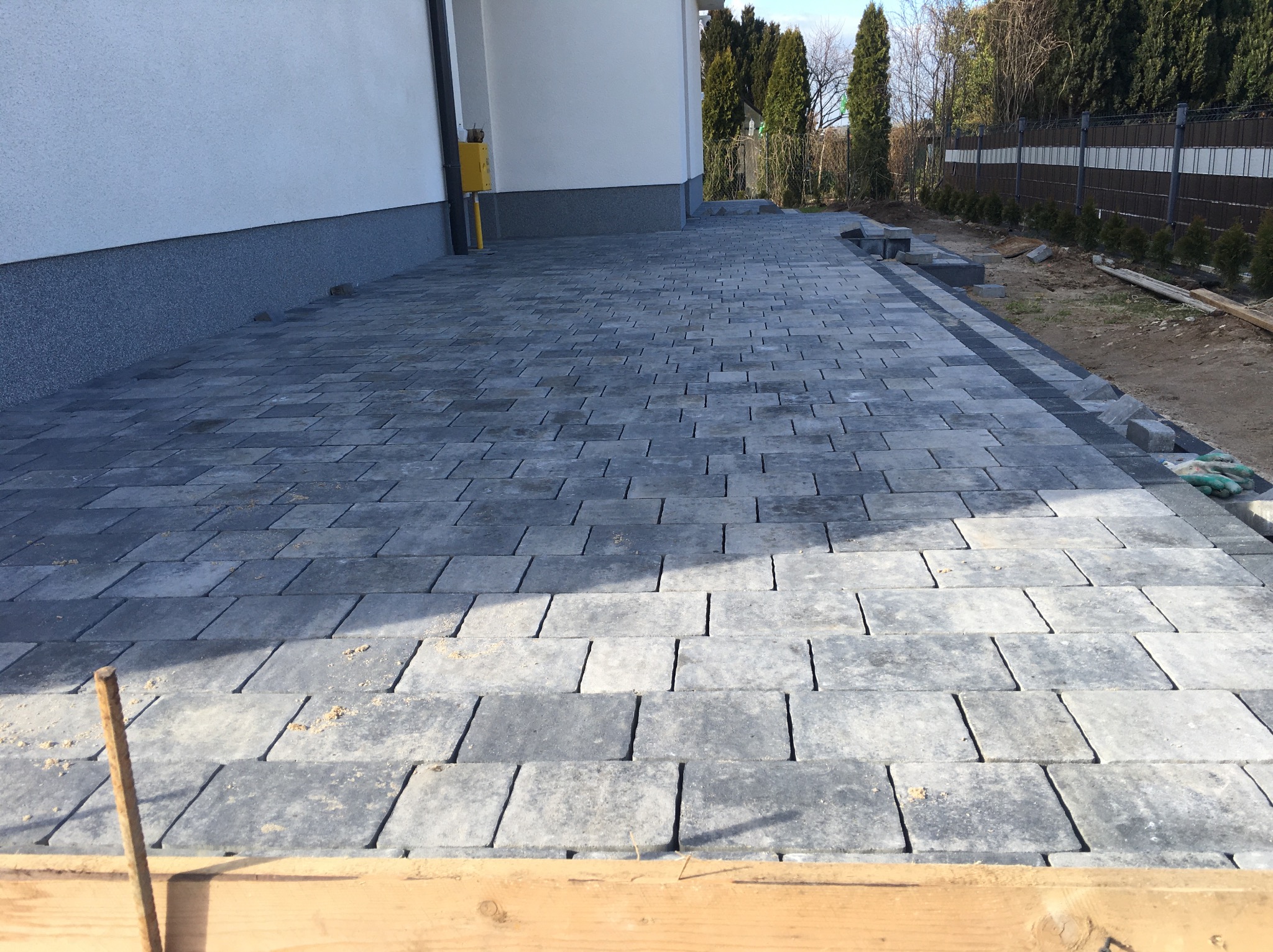 Ułożona szara kostka brukowa wzdłuż ściany budynku, widoczne resztki piasku i zabezpieczenie krawędzi deską, fragment ogrodu w tle.