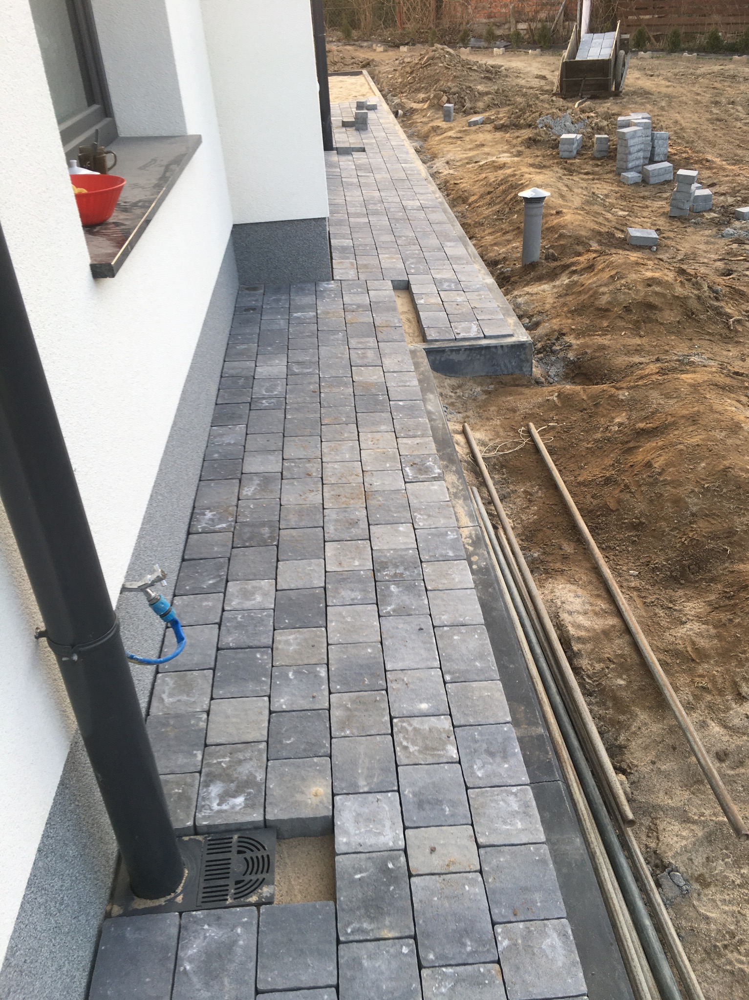 Niedokończona ścieżka z szarej kostki brukowej wzdłuż białej ściany budynku, widoczny odpływ liniowy, rury i przygotowany teren z kostką w oddali.