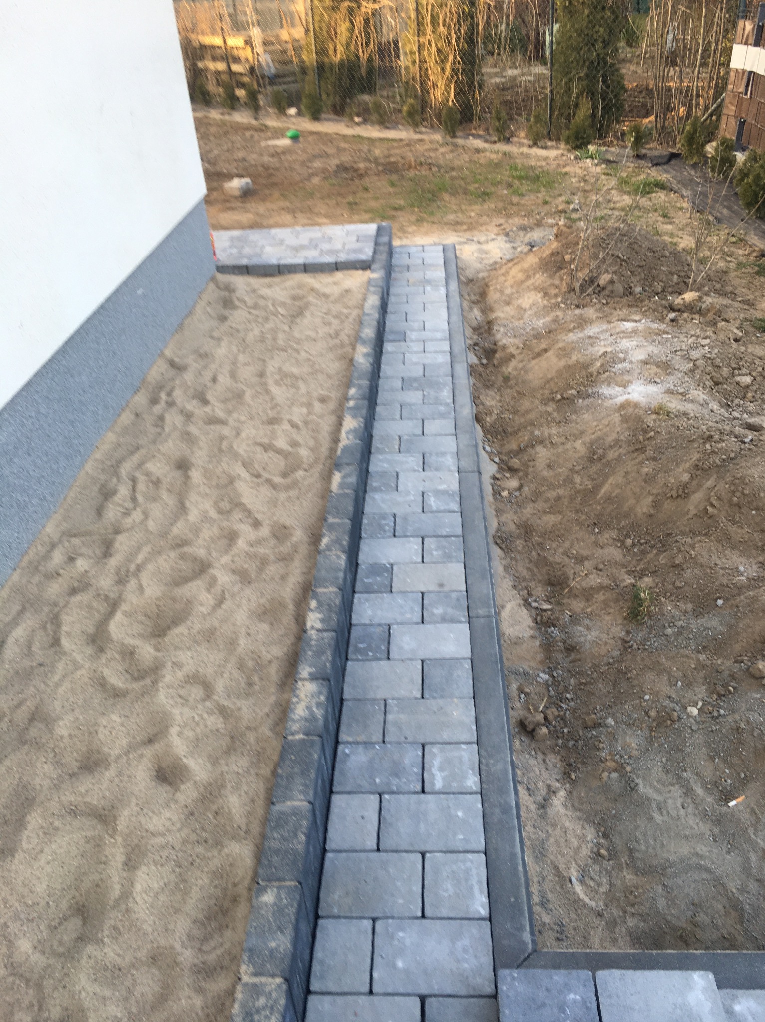 Świeżo ułożona ścieżka z szarej kostki brukowej obok budynku, widoczne przygotowane podłoże z piasku i ziemi.