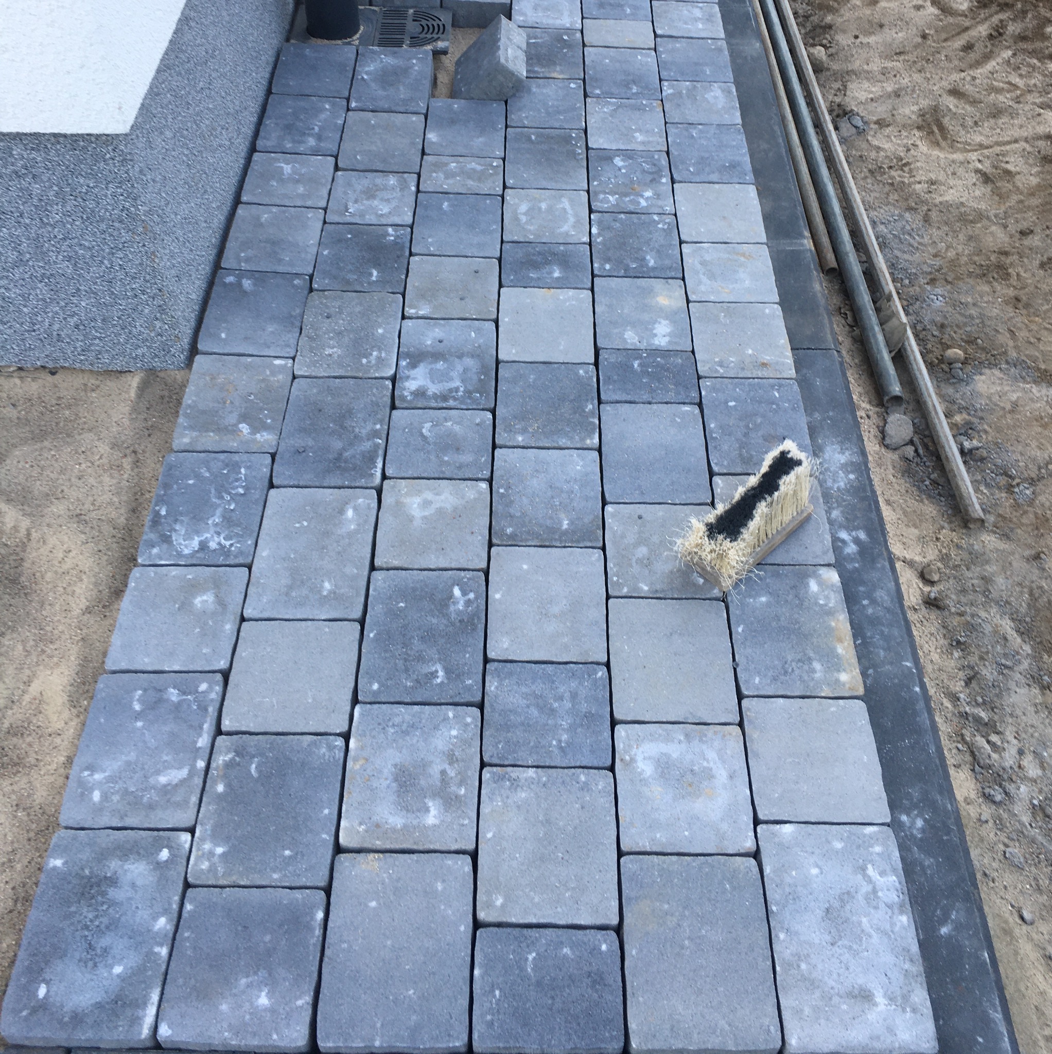 Fragment układanej szarej kostki brukowej, widoczny fragment krawężnika i szczotka do zamiatania fug, w tle ziemia.