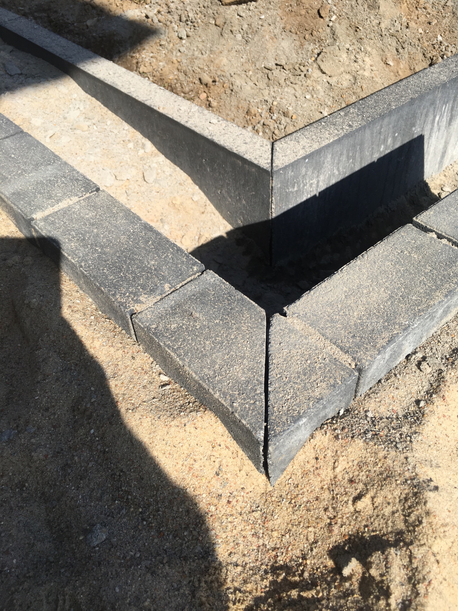 Krawężnik z szarej kostki betonowej ułożony na podsypce piaskowej, narożne połączenie elementów, cień rzucany na piasek.