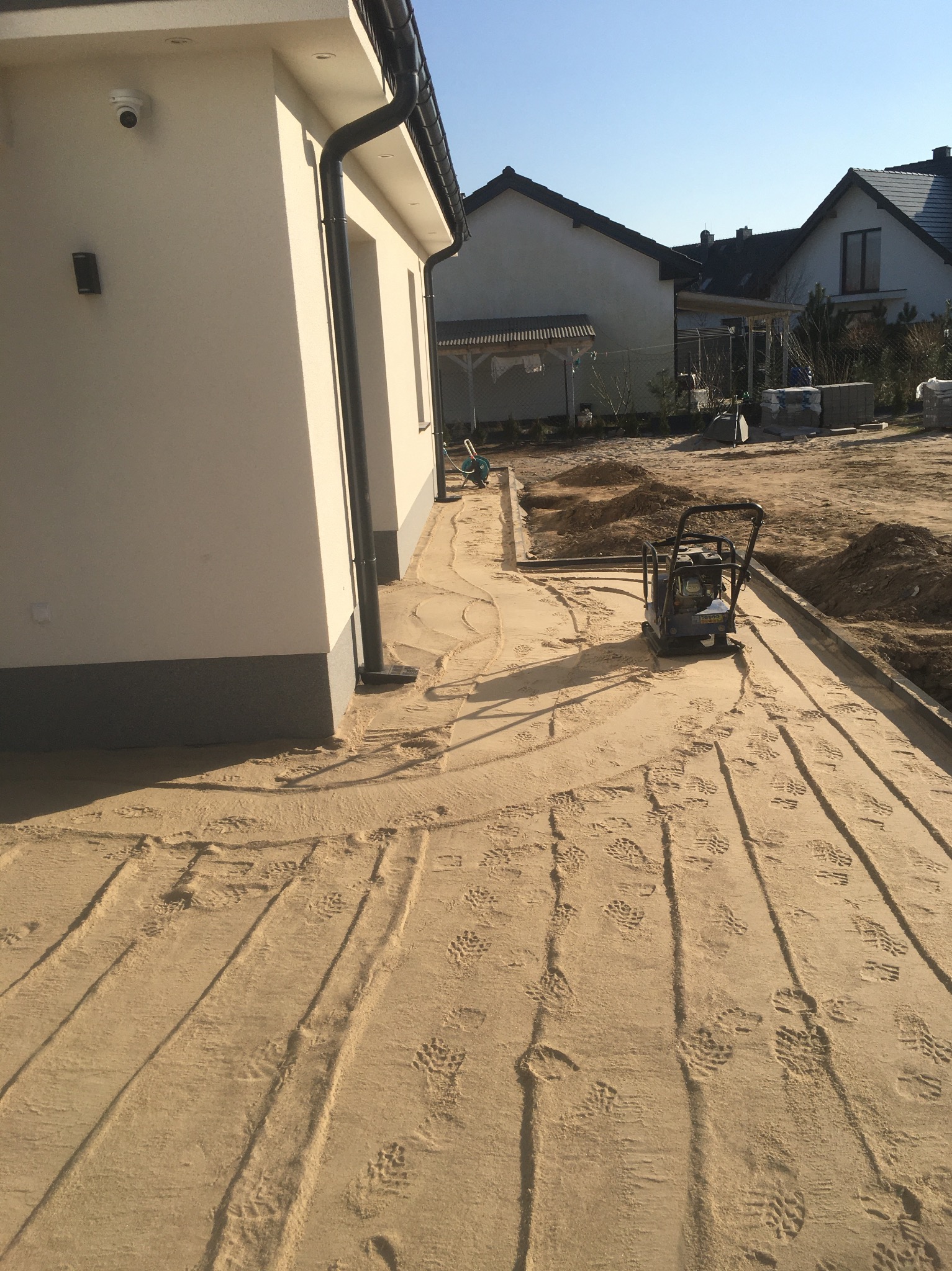 Przygotowanie podłoża pod kostkę brukową: ubijanie piasku zagęszczarką wzdłuż obrzeży, widoczne ślady butów i niedokończone wykopy.